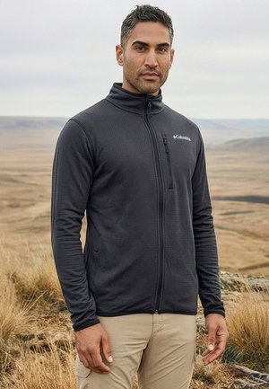 Mann, der eine schwarze Fleecejacke mit Reißverschluss und beige Hosen trägt, steht in einer trockenen, grasbewachsenen Landschaft mit sanften Hügeln im Hintergrund.