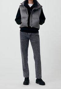 Persona che indossa un gilet imbottito grigio senza maniche sopra una felpa nera con cappuccio, jeans grigi slavati e stivali neri robusti, in piedi con le mani nelle tasche.