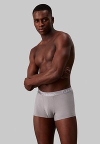 Sivi bokserice z gladko teksturo in širokim elastičnim pasom, na katerem je v beli barvi logo "Calvin Klein". Model stoji s prekrižanimi rokami.