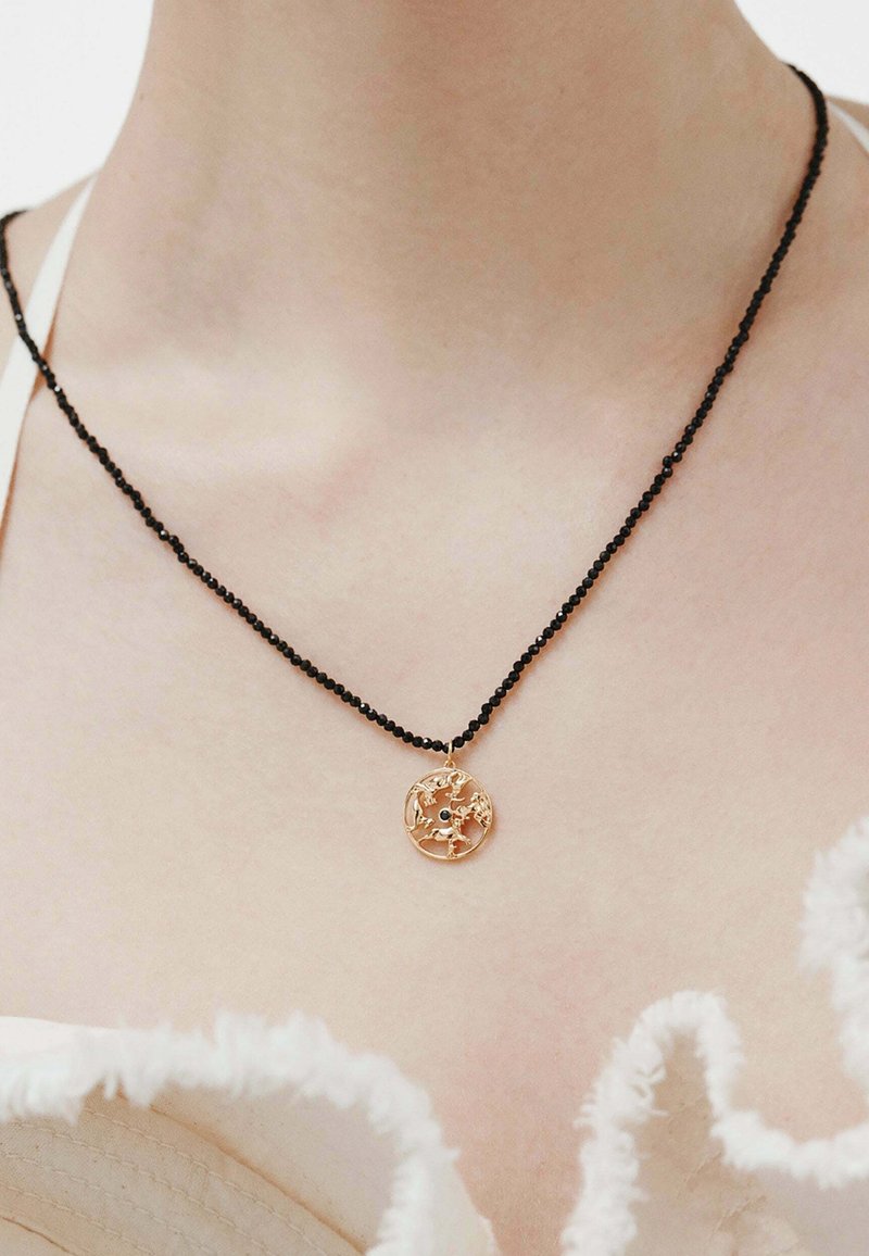 Collar de cuentas negras con un colgante circular dorado que presenta un diseño recortado intrincado y una piedra negra central.