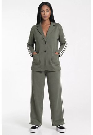 Kobieta ubrana w oliwkową marynarkę na dwa guziki i pasujące szerokie spodnie z białymi bocznymi paskami oraz czarne trampki, stoi z rękami w kieszeniach.