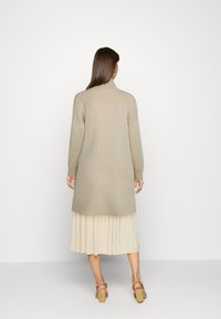 Beige dlouhý cardigan s vysokým límcem a dlouhými rukávy, s hladkou texturou, nošený přes plisovanou krémovou sukni a boty na blokovém podpatku.