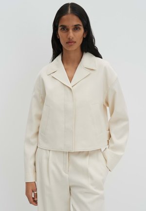 Jonge vrouw met lang donker haar, gekleed in een crèmekleurig op maat gemaakt jasje en bijpassende losse broek, staand tegen een effen lichte achtergrond.