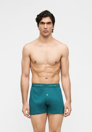 Homme torse nu aux cheveux foncés portant un boxer Calvin Klein turquoise, debout face à un fond clair uni.