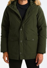 Olivgrüne Parka-Jacke mit kapuze, die mit Kunstfell gefüttert ist, Knopfverschluss, zwei Fronttaschen und einer strukturierten Stoffoberfläche.