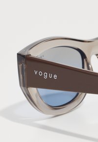 Óculos de sol de plástico castanho com uma armação translúcida, lentes azuis e a marca "vogue" em prateado na haste. Design elegante e angular.