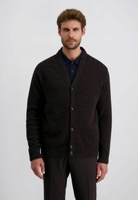 Man in een donker knooptrui over een marineblauw shirt en donkere broek, staand tegen een effen lichte achtergrond.