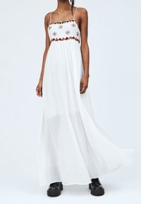 Weißes Maxi-Kleid mit einem floral bestickten Oberteil, dünnen Trägern und fließendem, durchsichtigem Stoff, kombiniert mit schwarzen klobigen Stiefeln.