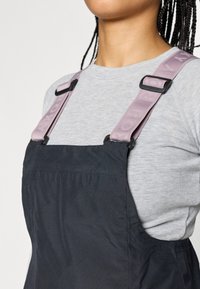 Macacão preto com tecido texturizado, apresentando alças ajustáveis cor-de-rosa claro com marca, usado sobre uma camisola de manga longa cinza.