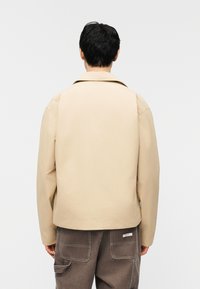 Άνδρας που κοιτάζει πίσω, φοράει μπεζ μπουφάν και σκούρα καφέ παντελόνια cargo με τσέπες-μπατζάκια, σε απλό λευκό φόντο.