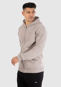 Lichtbeige zip-up hoodie met een voorzak en ribgebreide manchetten, gemaakt van zachte stof, met een klein logo op de borst. Draagt over zwarte broek.