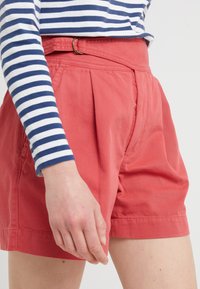 Pantaloncini plissettati color corallo con vita alta e passanti per cintura; abbinati a una maglietta a maniche lunghe a righe blu navy e bianche. Tessuto dalla texture liscia.