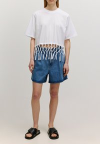 Witte crop top met een knoopfranje ontwerp aan de onderkant, gecombineerd met blauwe denim shorts en zwarte instapsandaal.