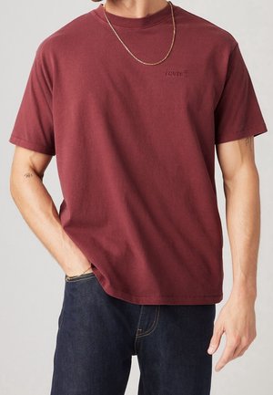 Mand iført en ensfarvet mørkerød Levi's t-shirt, mørkeblå jeans, guldkædehalskæde og små tatoveringer på begge underarme.