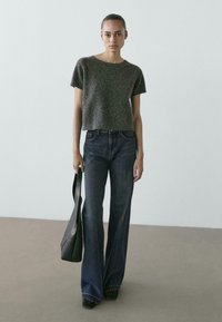 Młoda kobieta stojąca, ubrana w ciemnoszary sweter z krótkim rękawem, szerokie ciemne dżinsy, czarne buty i trzymająca czarną skórzaną torbę.