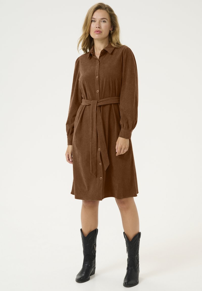 Robe chemise en velours côtelé marron avec un devant à boutons, des manches longues et une ceinture assortie à la taille. Portée avec des bottes de cowboy noires.