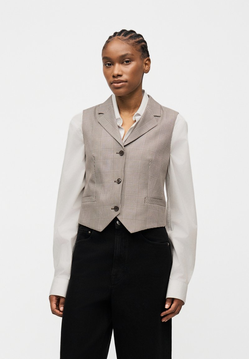 Γυναίκα με πλεξούδες, φορούν μπεζ καρό αμάνικο γιλέκο με κουμπιά πάνω από λευκό πουκάμισο μακρυμάνικο και μαύρο παντελόνι ψηλόμεσο, στέκεται μπροστά σε λευκό φόντο.