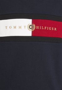 Jersey de algodón azul marino con rayas horizontales en negro, blanco y rojo, y detalles del logo y emblema en dorado "TOMMY HILFIGER".