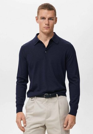 Polo à manches longues bleu marine avec une texture douce, un col classique et trois boutons. Porté avec un pantalon de couleur claire et une ceinture noire.