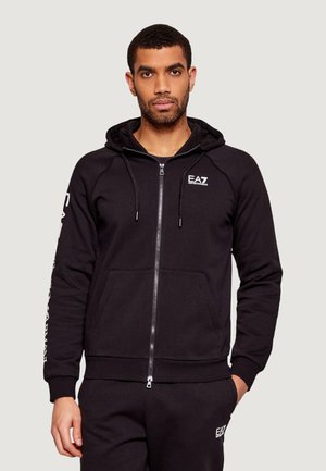 Uomo che indossa felpa nera con zip e pantaloni neri coordinati con logo EA7 Emporio Armani sul petto e sulla coscia sinistra, in piedi con una mano in tasca.