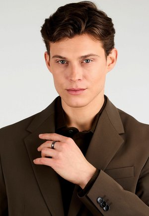 Blazer marrone con rever a punta, indossato sopra una camicia scura. Mano posizionata vicino al viso, con una fascia d'argento visibile. Tre bottoni neri sul polsino.