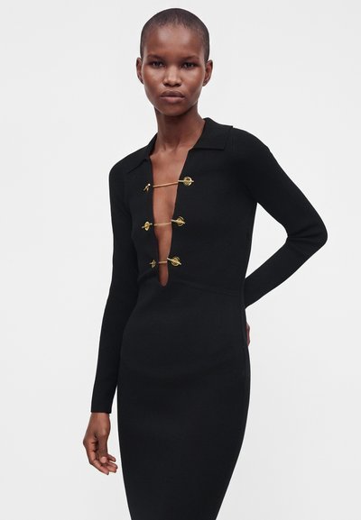 Femme portant une robe noire à manches longues avec un profond décolleté en V maintenu par trois fermoirs à bascule dorés.