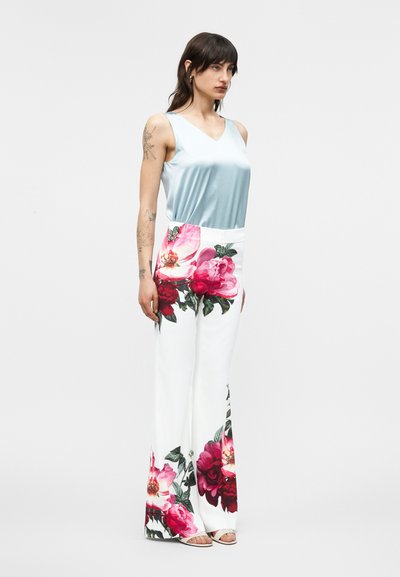 Mujer con top de satén sin mangas de color azul claro y pantalones anchos blancos con grandes estampados florales en rosa y rojo, de pie frente a un fondo liso.