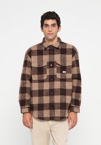 Bruin en beige geruite flanellen shirt met een knoopsluiting aan de voorkant, twee borstzakken en een label aan de linkerkant. Zachte textuur met een losse pasvorm.