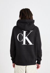Sudadera con capucha negra con un gran logo blanco "cK" en la espalda. Presenta una textura suave y un diseño de ajuste holgado.