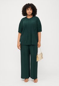 Une femme aux cheveux bouclés porte un haut plissé vert et un pantalon large assorti, tenant un sac à main doré, se tenant devant un fond blanc.