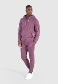 Uomo in piedi che indossa un completo coordinato di felpa viola e pantaloni sportivi, con sneakers bianche, mani nelle tasche, che guarda leggermente a sinistra su uno sfondo neutro.