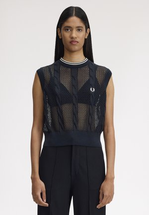 Femme aux longs cheveux foncés portant un haut noir sans manches en tricot avec des motifs torsadés par-dessus un soutien-gorge noir et un pantalon noir taille haute.