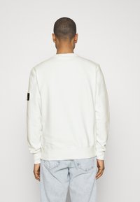 Hanorac alb cu manșete și tiv ribbed. Prezintă un mic patch cu logo negru pe mâneca stângă. Textură netedă și design clasic de guler rotund.