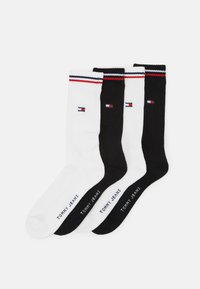 Tres pares de calcetines de tripulación: uno blanco, uno negro, ambos con textura acanalada y acentos en rojo, blanco y azul en la parte superior. "TOMMY JEANS" impreso en las suelas.