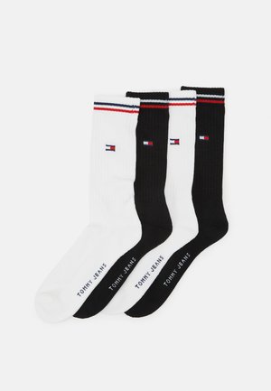 Três pares de meias de crew: uma branca, uma preta, ambas com textura canelada, e detalhes em riscas vermelhas, brancas e azuis na parte de cima. "TOMMY JEANS" impresso nas solas.