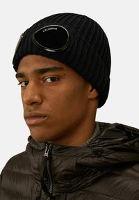 Beanie nero lavorato a maglia con texture a costine, dotato di occhiali riflettenti sulla parte anteriore. Indossato con un giubbotto gonfio scuro per tenere caldo.