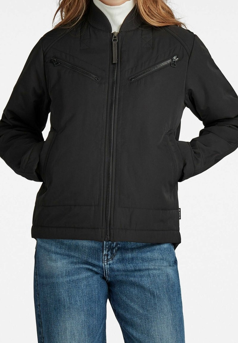 Czarna bomberka z zamkiem z przodu, dwoma kieszeniami na zamek na piersi, żebrowanym kołnierzem i ciepłym, teksturowanym materiałem. Nosi się ją na niebieskich dżinsach.