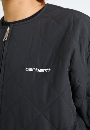 Person iført en sort quiltet jakke med frontlynlås og hvidt broderet Carhartt-logo på brystet.