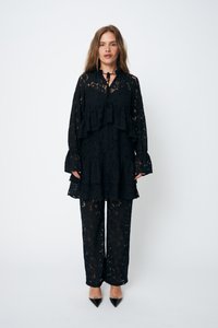 Abito in pizzo nero caratterizzato da una tunica a strati con un collo alto e maniche a ruffle, abbinato a pantaloni in pizzo coordinati e tacchi a punta.