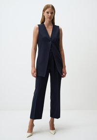 Completo gilet blu navy con scollatura a V profonda e chiusura a singolo bottone, abbinato a pantaloni dritti e décolleté bianche appuntite. Texture liscia.
