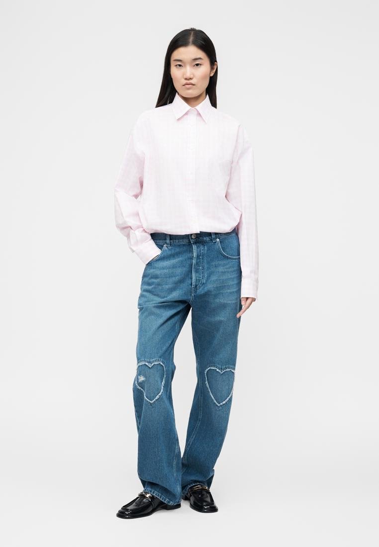 Femme portant une chemise rose clair rentrée dans un jean bleu avec des patchs en forme de cœur aux genoux et des mocassins noirs, debout devant un fond blanc uni.