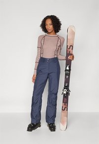 Slidinėjimo apranga, kurios sudėtyje yra rožinės ir kreminės spalvos ilgomis rankovėmis margintas viršus, derinamas su tamsiai mėlynomis vandeniui atspariomis kelnėmis. Slidinėjimo įranga apima juodus batus ir slides.