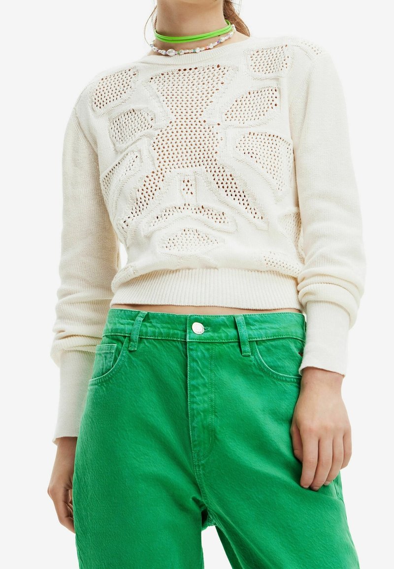 Pull blanc en maille avec un motif ajouré géométrique, associé à un pantalon taille haute vert vif et des colliers superposés verts et perles.