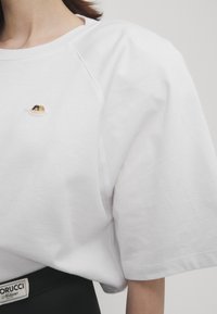 White cotton t-shirt with short, raglan sleeves. Features a small embroidered detail on the chest. Soft texture, relaxed fit, minimalistic design.