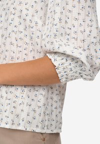 Witte blouse met kleine blauwe en bruine bloemenpatronen, gestructureerde stof en geplooide mouwen. Verticale strepen versterken het ontwerp.