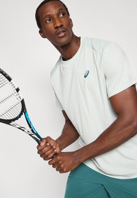 Αθλητική μπλούζα ανοιχτού γκρι χρώματος με κοντά μανίκια, που φέρει μικρό λογότυπο. Το μοντέλο κρατάει μια μαύρη ρακέτα τένις με μπλε λεπτομέρειες, σε απλό φόντο.