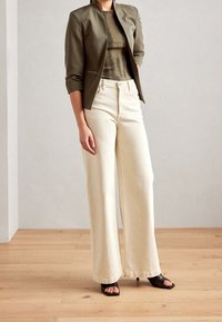 Jasnozielona dopasowana kurtka z detalem na zamek, zestawiona z jasnobeżowymi spodniami o szerokich nogawkach z wysokim stanem oraz czarnymi sandałami na obcasie, na drewnianej podłodze.