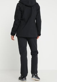 Black waterproof outdoor jacket with hood, textured fabric, and adjustable cuffs, paired with matching black trousers and athletic shoes.