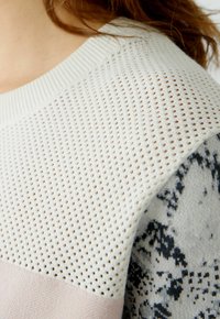 Strickpullover mit einer cremefarbenen, durchbrochenen Textur oben, einem hellrosa, einfarbigen Abschnitt darunter und grauen Musterakzenten, die die geschichtete Bauweise präsentieren.