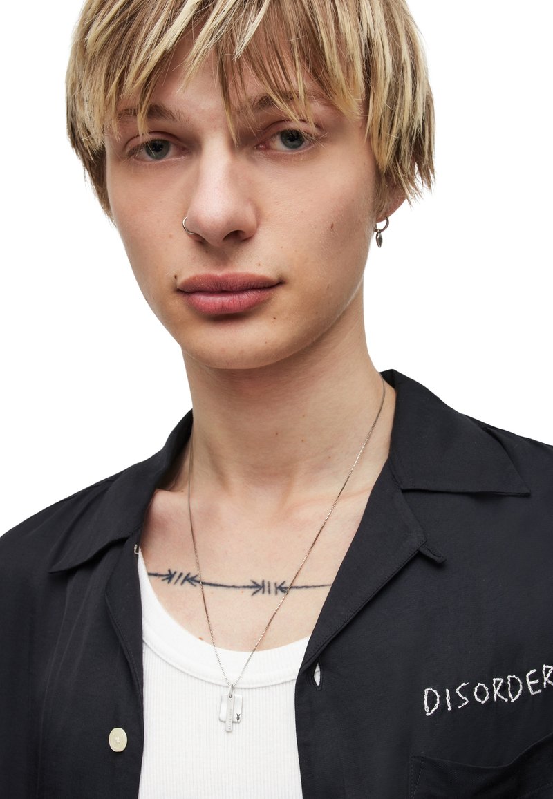 Sort kortærmet skjorte med broderet "DISORDER" og en hvid tanktop under; lagdelte sølvhalskæder; synlig tatovering med pilmotiver.